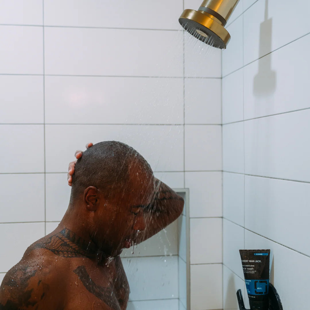 Person showering with a shower head and a tube of shampoo or conditioner on a tiled wall.